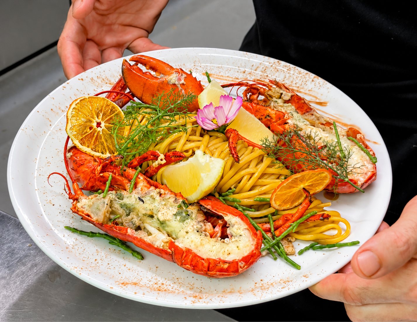 Deux mains tiennent une assiette blanche garnie de homard grillé, de spaghettis, de rondelles d'orange séchée, de citron, de fenouil et d'une fleur rose.