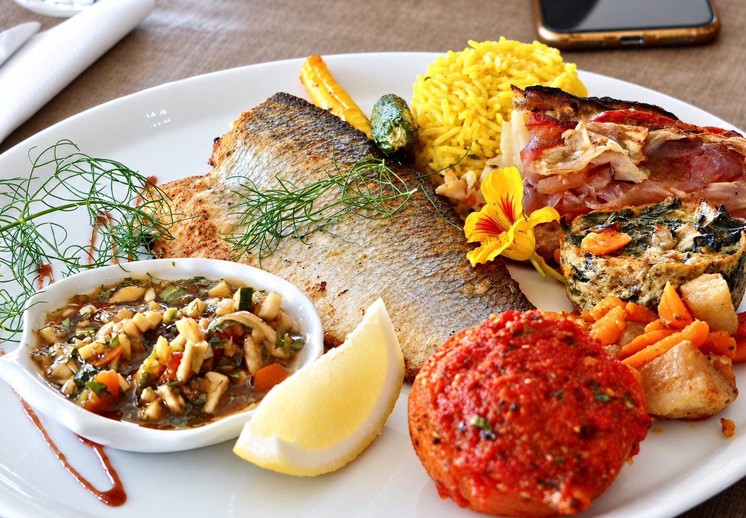 Une assiette blanche garnie d'un filet de poisson grillé, de riz jaune, de tomate farcie, de légumes rôtis, d'une sauce en coupelle, d'un quartier de citron et d'une fleur de capucine.