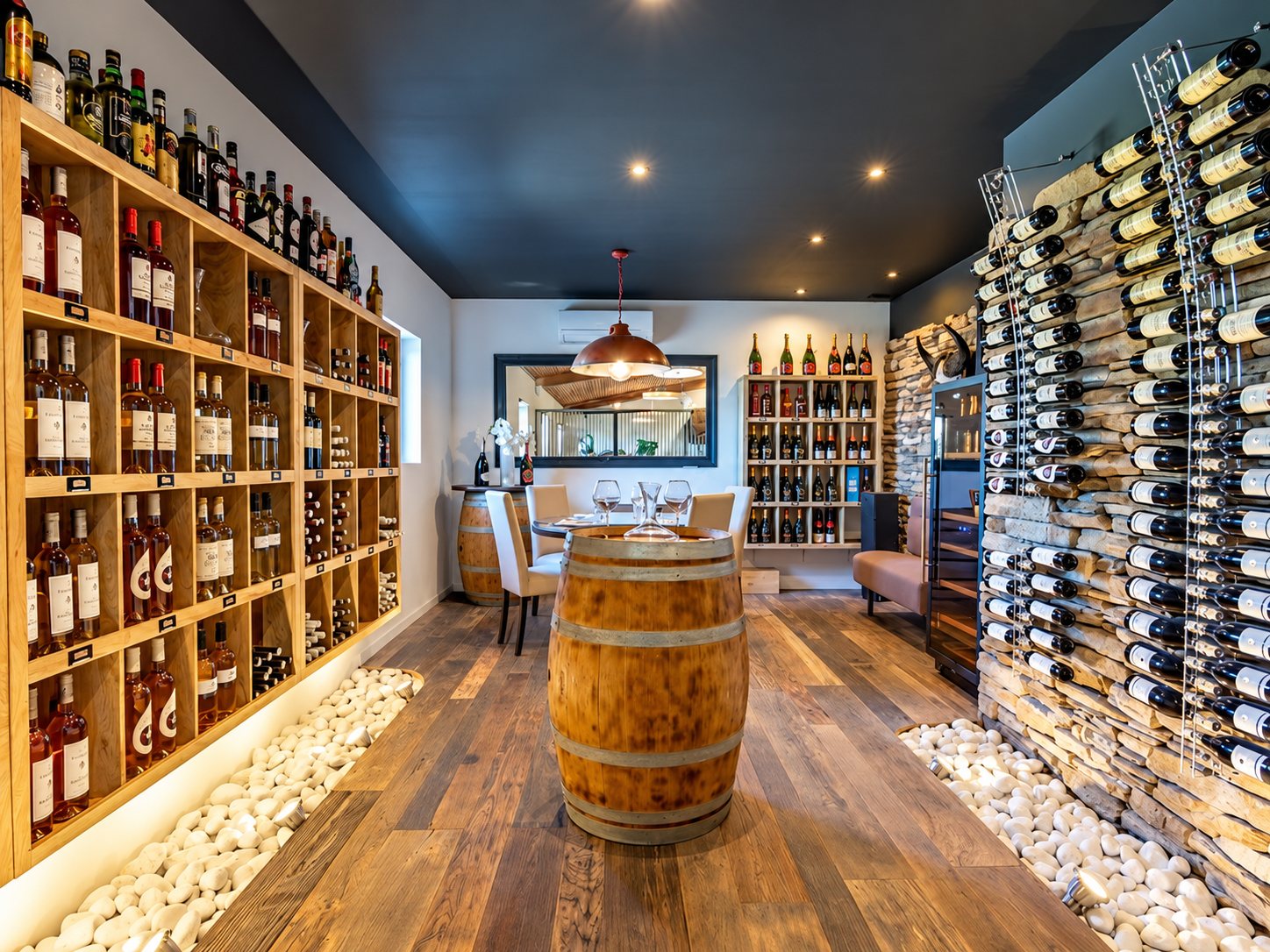 Intérieur d'une cave à vin avec étagères en bois remplies de bouteilles, un tonneau au centre, une table dressée au fond et un mur de pierre qui expose des bouteilles couchées.