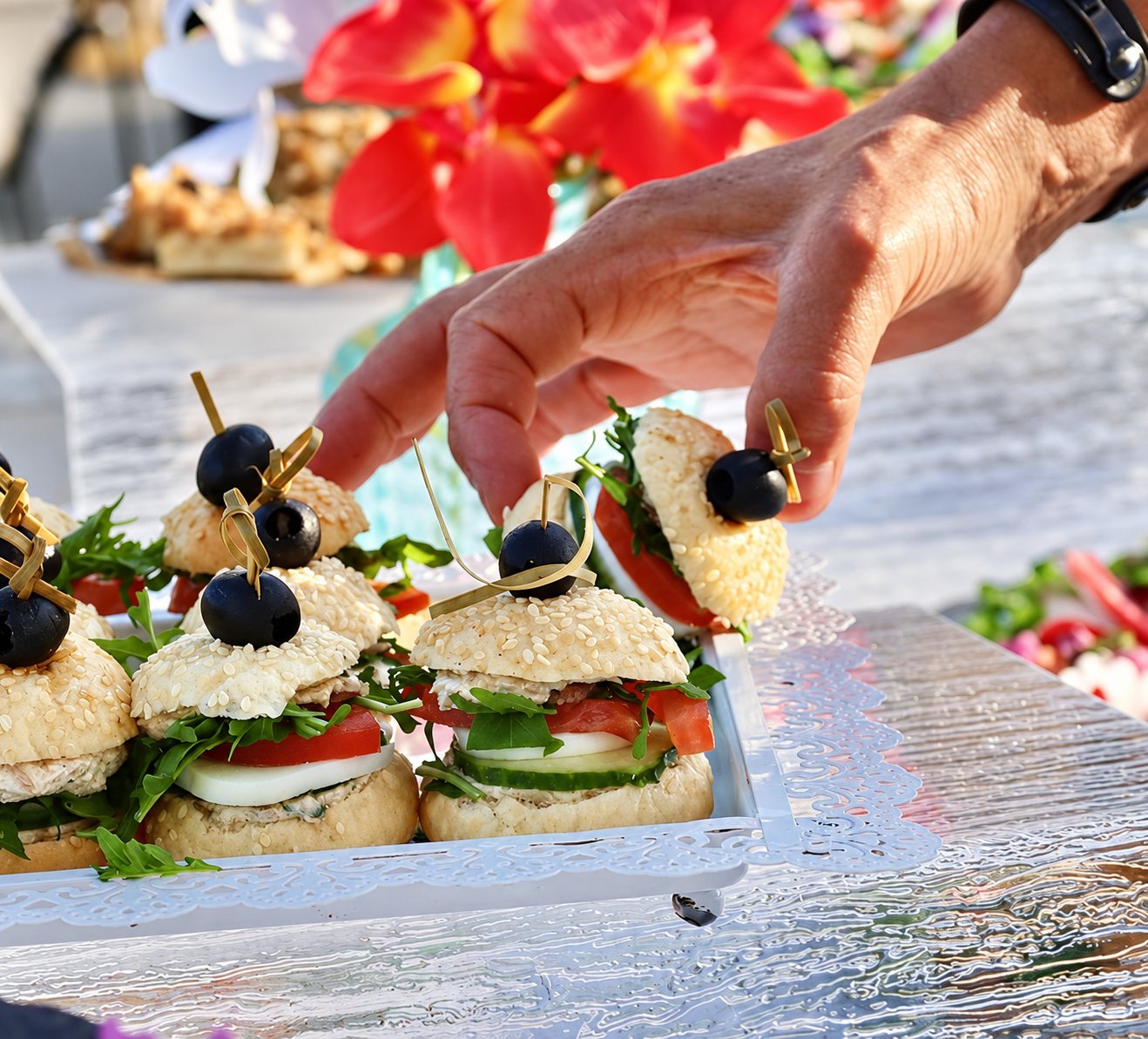 Une main saisit un petit burger garni d'olive noire, de tomate, de concombre et de roquette, posé sur un plateau blanc ajouré lors d'un buffet en extérieur.