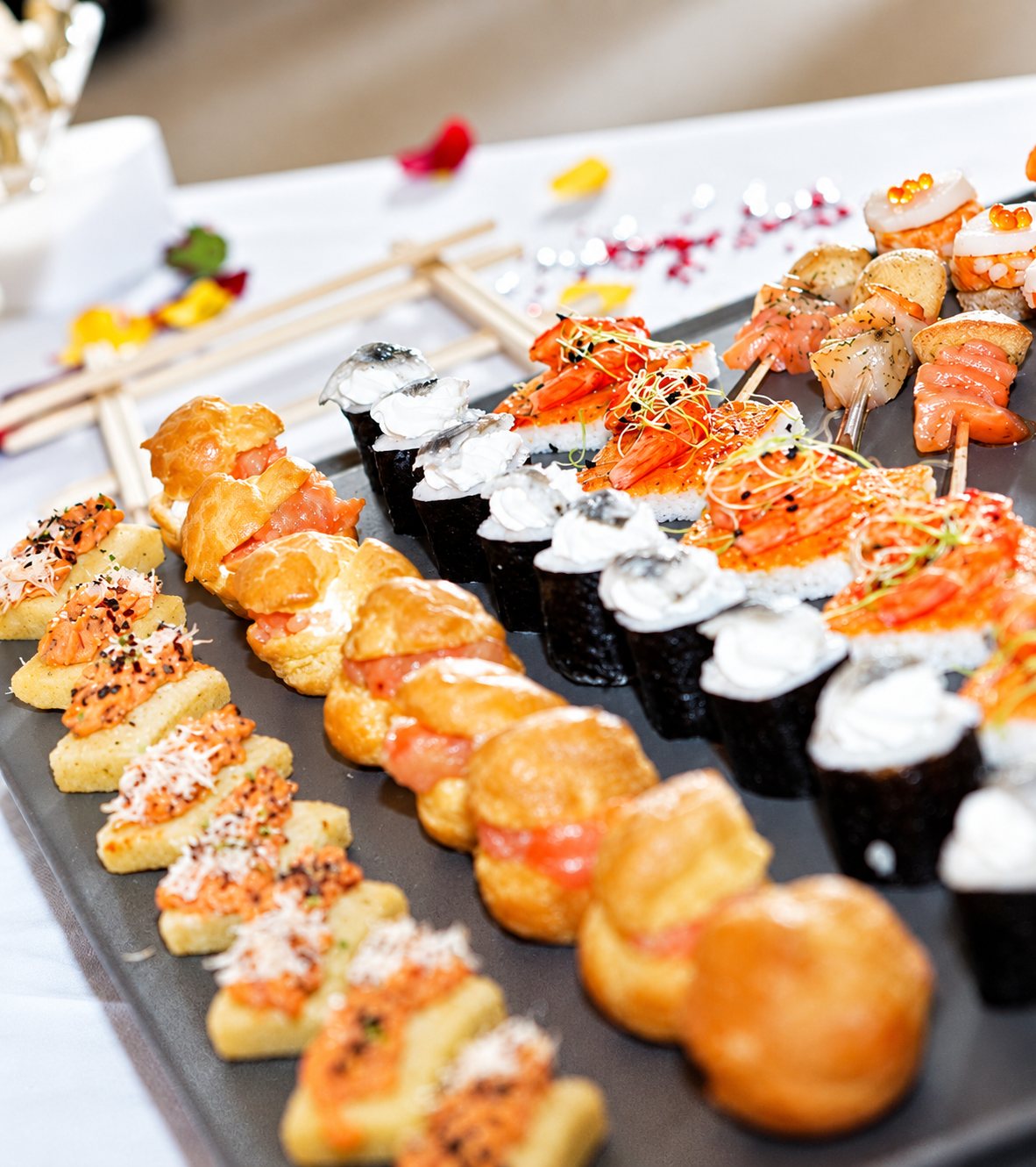 Un plateau gris garni de canapés, de petits choux, de makis et de brochettes au saumon, posé sur une table dressée avec des baguettes.