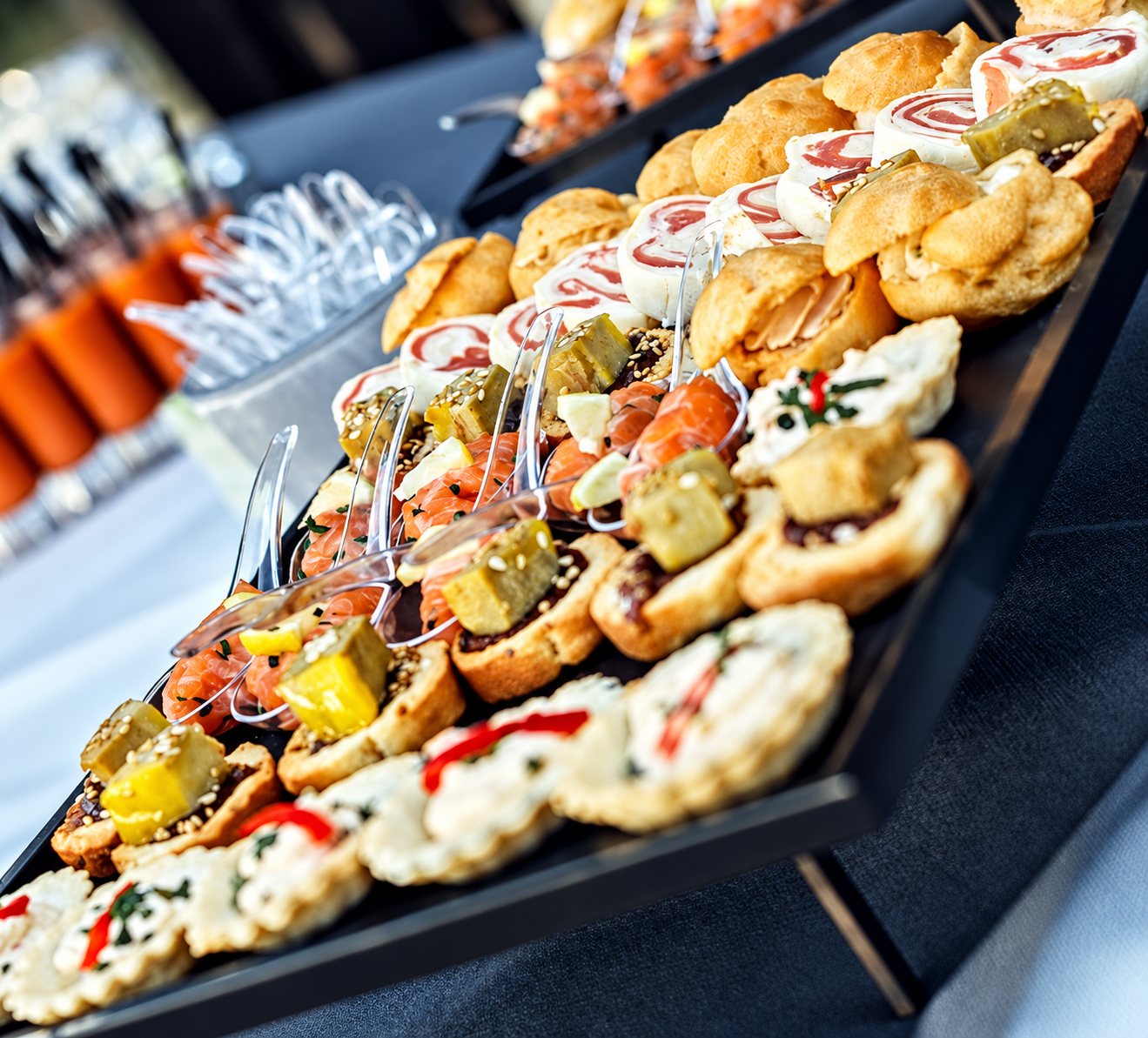 Des plateaux noirs garnis de canapés, mignardises salées, roulés au saumon et petits choux sont disposés sur un buffet, avec des verrines orangées en arrière-plan.