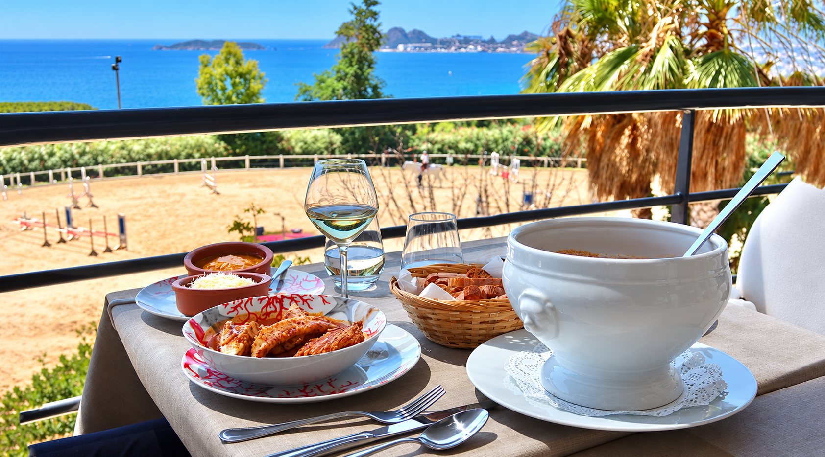 Sur une table de terrasse, une soupière, un plat de raviolis, un verre de vin blanc et un panier de pain font face à la mer et à un manège d'équitation.