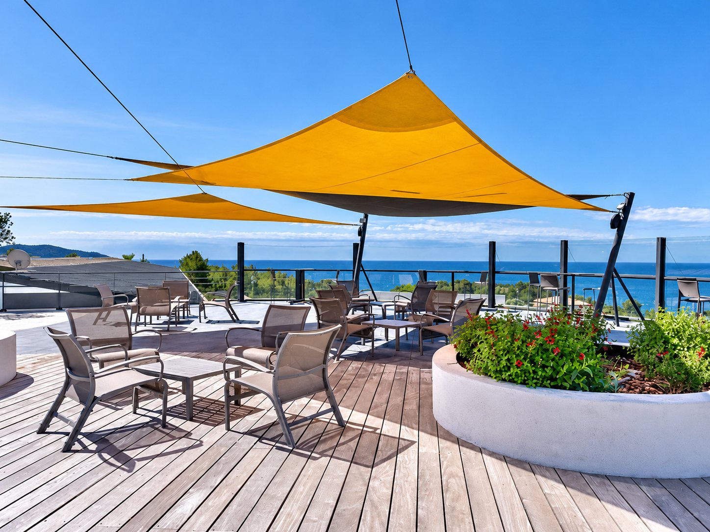 Une terrasse en bois meublée de fauteuils et de tables basses, abritée par des voiles d'ombrage jaunes, avec la mer en arrière-plan.