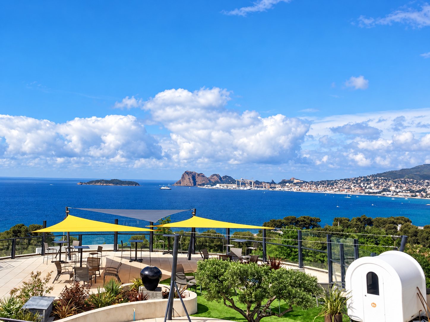 Une terrasse aménagée avec tables, chaises et voiles d'ombrage jaunes offre une vue sur la mer, une île et une ville côtière sous un ciel bleu.