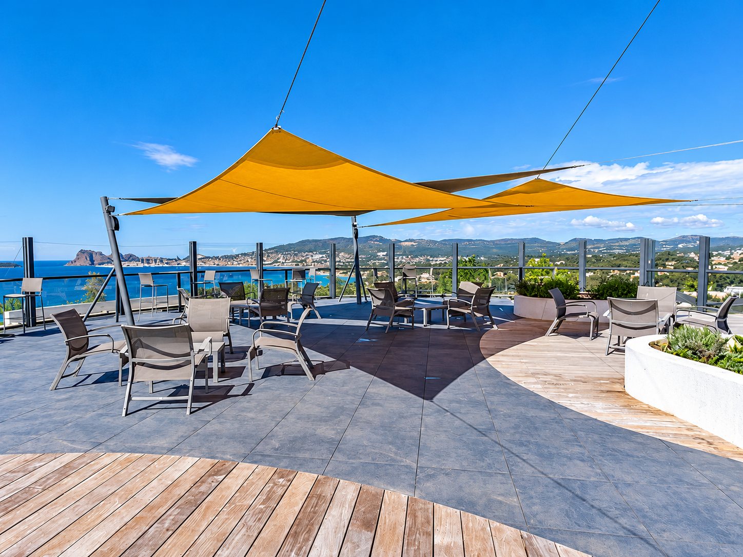 Une terrasse en hauteur aménagée avec des fauteuils et des tables sous des voiles d'ombrage orange, face à la mer et aux collines.
