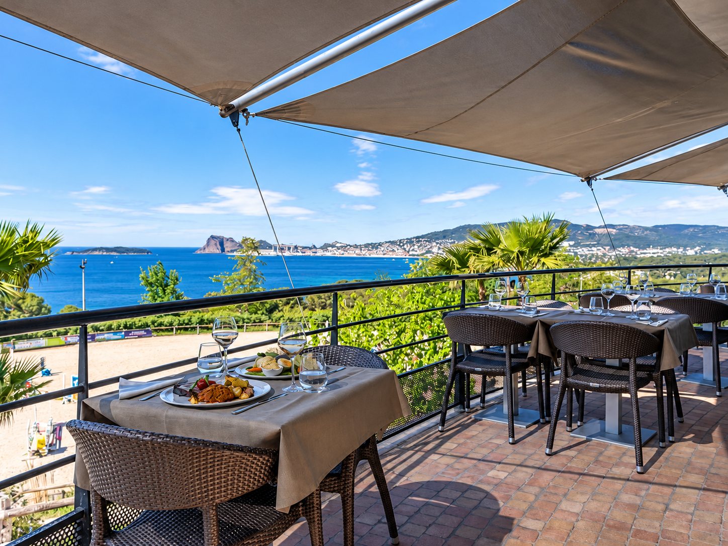 Des tables dressées avec assiettes et verres sur une terrasse ombragée par des voiles, face à la mer et à une baie bordée de collines.